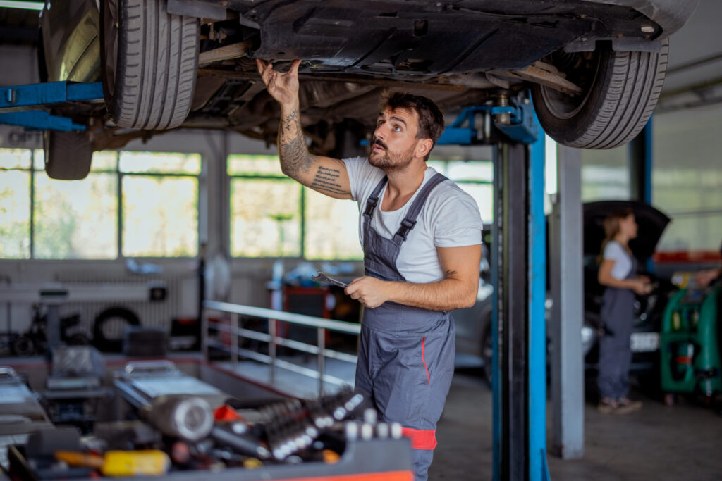 A man in overalls works on a car that is lifted in an auto mechanic shop.