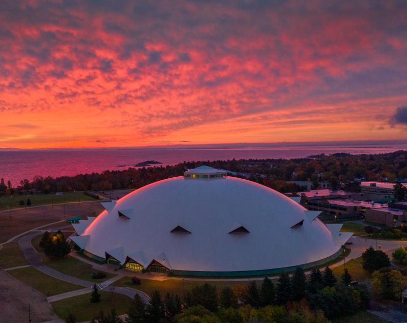 A view of the Superior Dome in Marquette, Michigan with a picturesque orange and pink sunrise in the background.