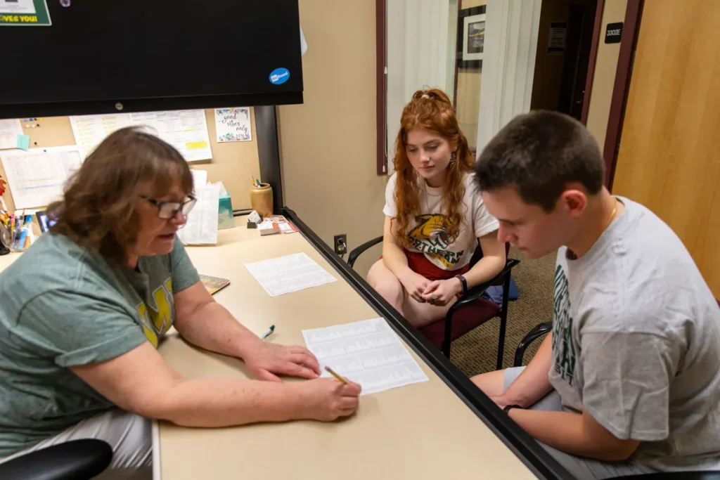 A woman at a desk sharing information with two NMU students wearing wildcat t-shirts.