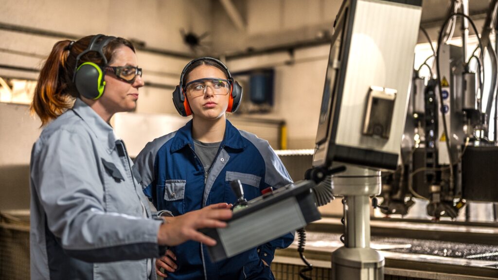 A skilled mature Caucasian woman operates a water cutting CNC machine, guiding her young female apprentice in protective gear within the bustling factory environment.