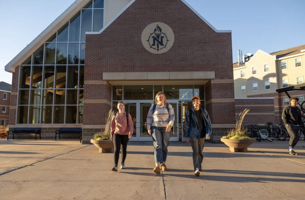 Three students walking out of a large brick building with the NMU torch logo on the front of the building.
