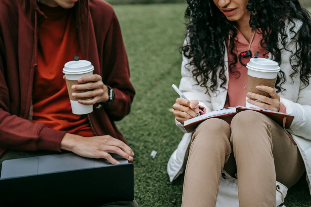 Unrecognizable students with takeaway beverage browsing laptop and writing in copybook while doing homework together on grassy ground on street